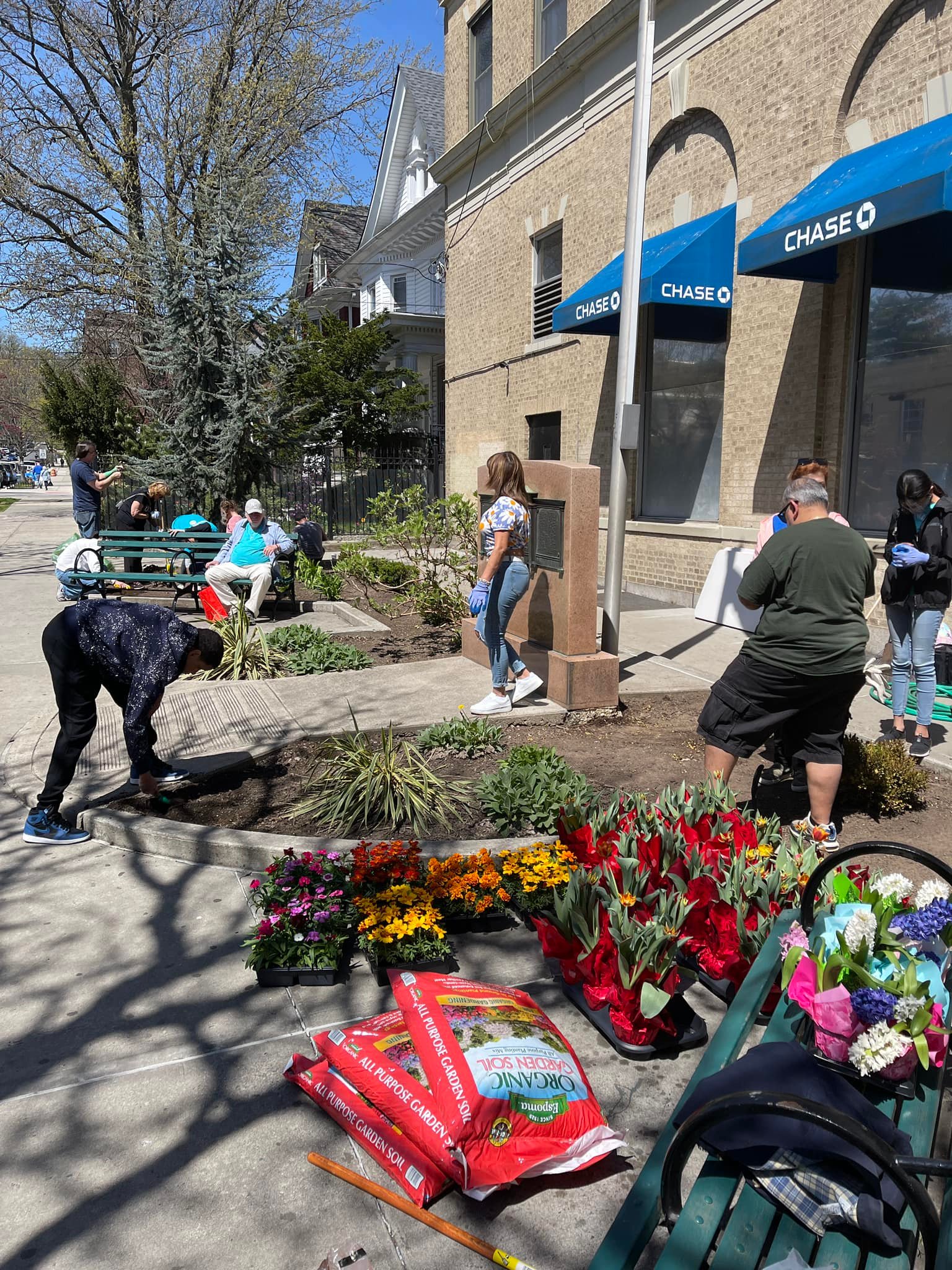 People participate in a community gardening event on a sunny day, planting flowers and working with soil near a Chase bank. Flower pots, soil bags, and blooming plants are arranged on the sidewalk.