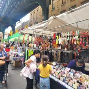 Outdoor street market scene under an elevated train track, with people browsing colorful stalls selling wallets, accessories, and hanging decorations. Vendors and shoppers are engaged, and buildings line the background.