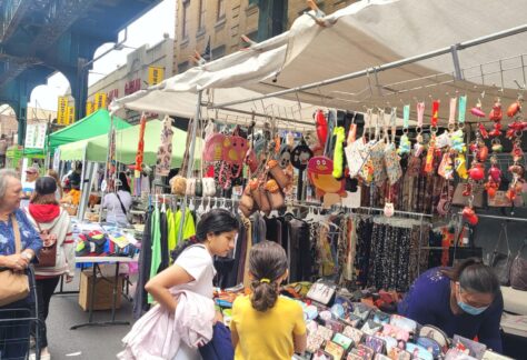 Outdoor street market scene under an elevated train track, with people browsing colorful stalls selling wallets, accessories, and hanging decorations. Vendors and shoppers are engaged, and buildings line the background.