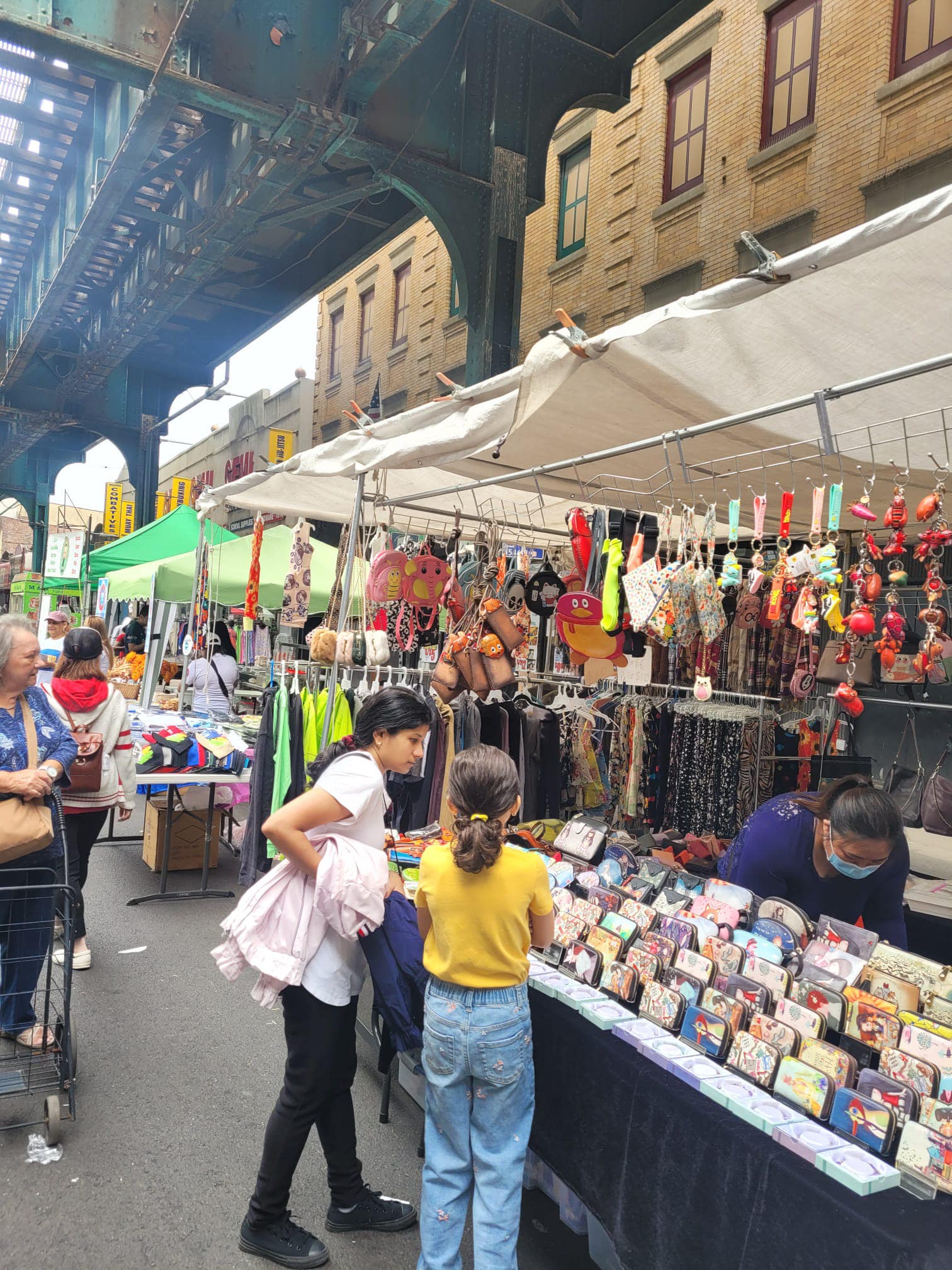 Outdoor street market scene under an elevated train track, with people browsing colorful stalls selling wallets, accessories, and hanging decorations. Vendors and shoppers are engaged, and buildings line the background.