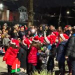 A group of children wearing Santa hats stand together outdoors at night, singing to a large crowd of bundled-up adults, many of whom are also wearing festive hats and taking photos.