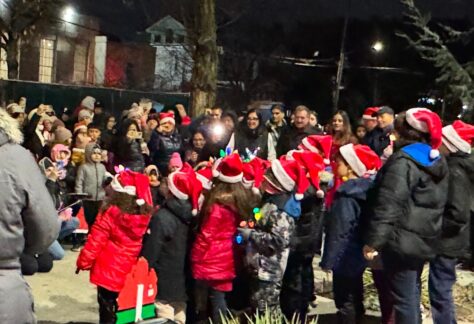 A group of children wearing Santa hats stand together outdoors at night, singing to a large crowd of bundled-up adults, many of whom are also wearing festive hats and taking photos.
