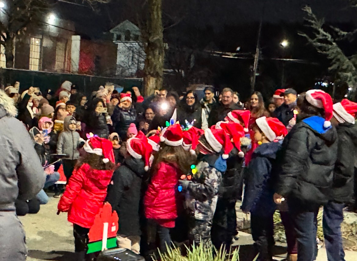 A group of children wearing Santa hats stand together outdoors at night, singing to a large crowd of bundled-up adults, many of whom are also wearing festive hats and taking photos.