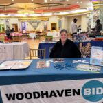 A woman sits behind a table covered with a "WOODHAVEN BID" banner at an indoor event. The table displays brochures, pens, and flyers. Other booths and people are visible in the background.