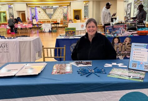 A woman sits behind a table covered with a "WOODHAVEN BID" banner at an indoor event. The table displays brochures, pens, and flyers. Other booths and people are visible in the background.