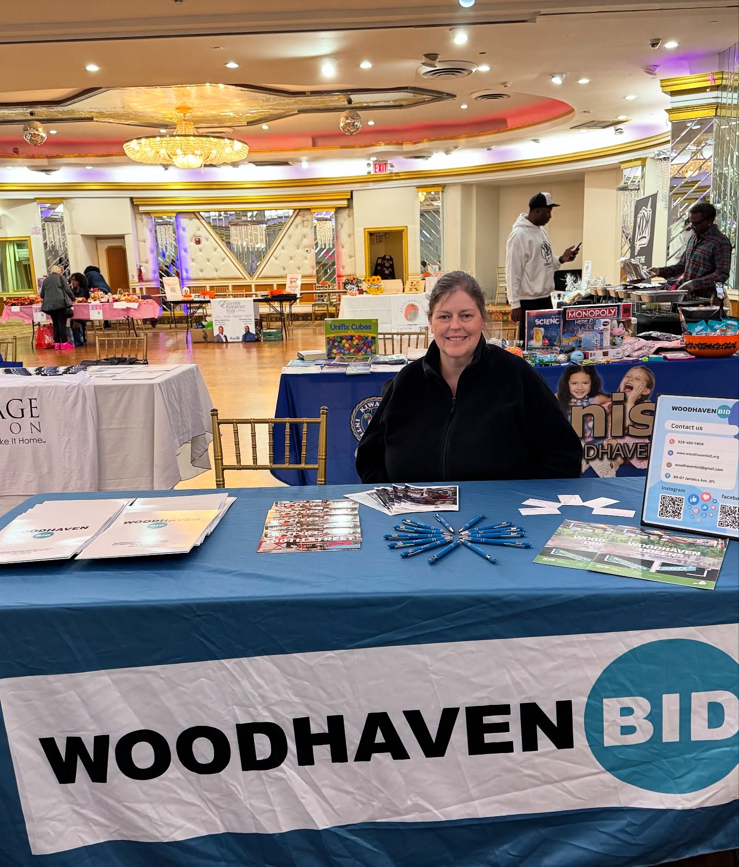A woman sits behind a table covered with a "WOODHAVEN BID" banner at an indoor event. The table displays brochures, pens, and flyers. Other booths and people are visible in the background.