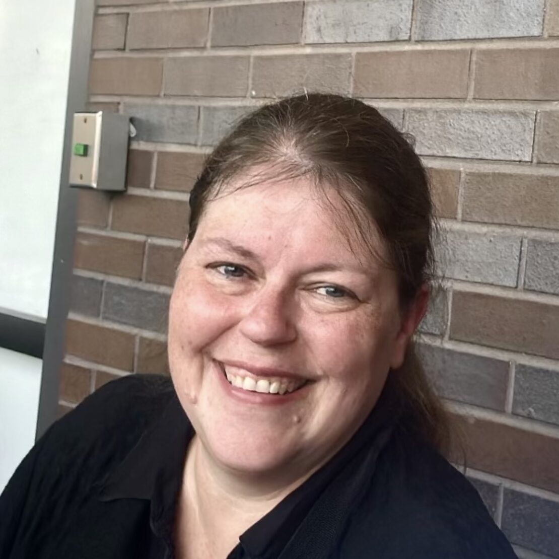 A woman with long brown hair pulled back smiles at the camera. She is wearing a black top and sits in front of a brick wall indoors.