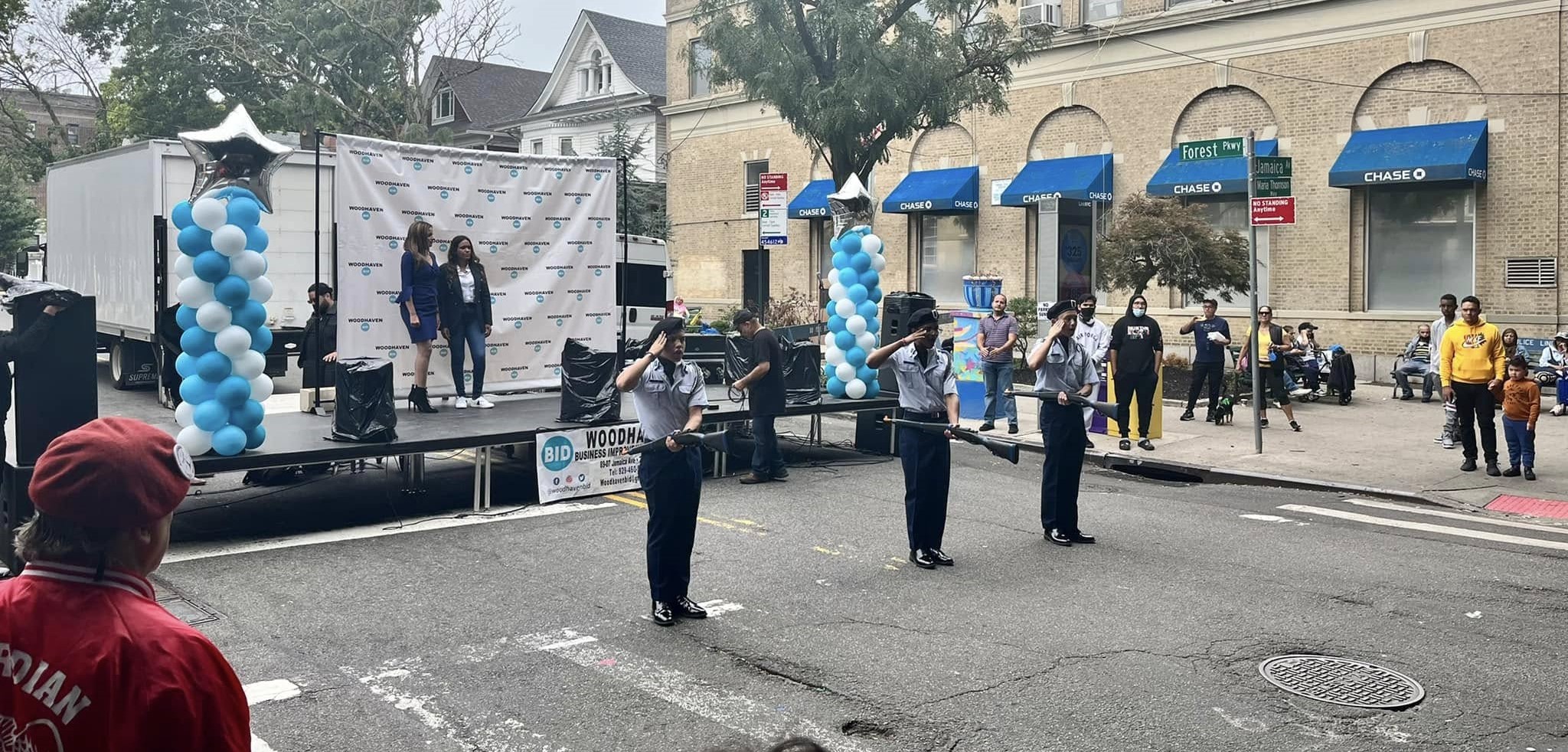 Three uniformed individuals perform a flag routine on a street, while two people stand on a stage with blue and white balloons. Bystanders watch from the sidewalk near a Chase bank.