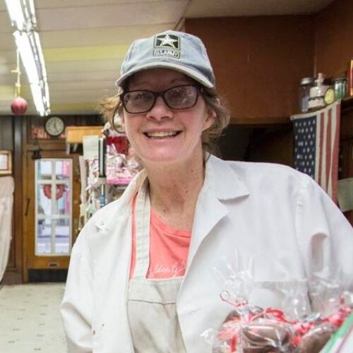A smiling woman wearing glasses, a cap, and a white coat stands in a shop next to a tray of chocolate-covered strawberries. The shop has a patriotic theme with an American flag in the background.