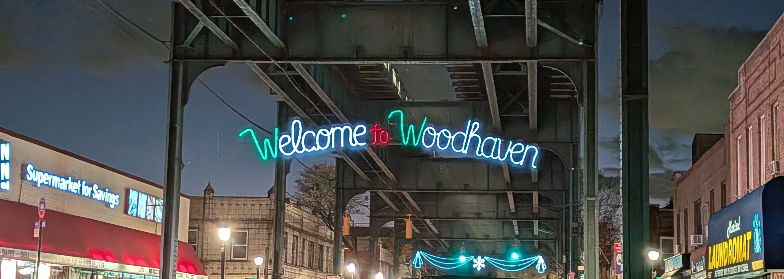 A neon sign reading "Welcome to Woodhaven" hangs under an elevated train track at night, with storefronts and streetlights illuminating the surrounding area.