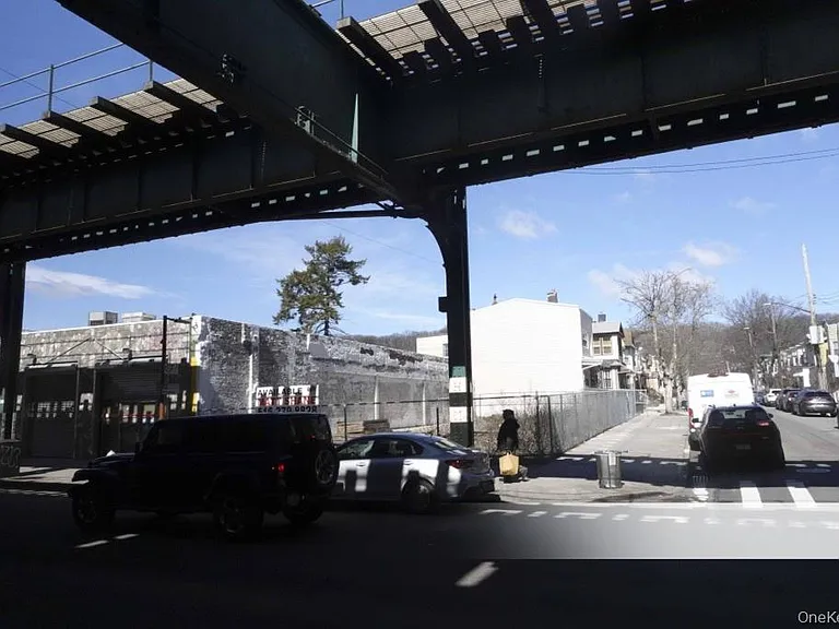 A city street scene beneath an elevated train track, with cars parked along the road and a person walking on the sidewalk. Buildings and trees are visible in the background under a clear blue sky.