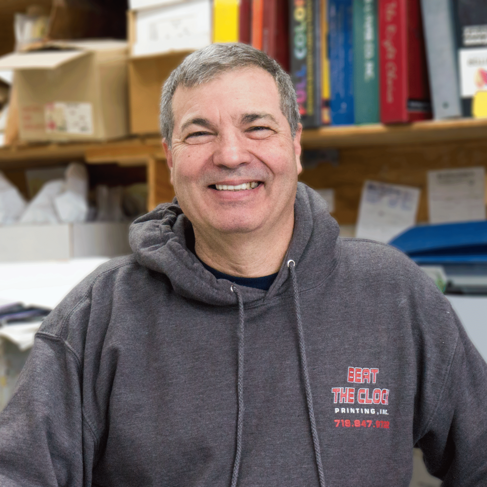 A smiling middle-aged man in a gray hoodie stands in a cluttered office or workshop with shelves of boxes, papers, and binders behind him. The hoodie reads ``Beat The Clog Printing, Inc.`` in red and white text.