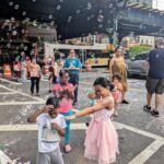Children and adults enjoy a lively street scene filled with bubbles. A girl in a pink dress playfully points a bubble wand at a smiling boy, while others watch and play under an elevated train track on a sunny day.