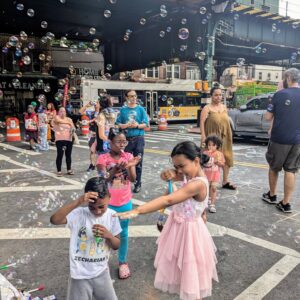 Children and adults enjoy a lively street scene filled with bubbles. A girl in a pink dress playfully points a bubble wand at a smiling boy, while others watch and play under an elevated train track on a sunny day.