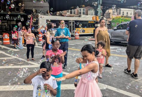 Children and adults enjoy a lively street scene filled with bubbles. A girl in a pink dress playfully points a bubble wand at a smiling boy, while others watch and play under an elevated train track on a sunny day.