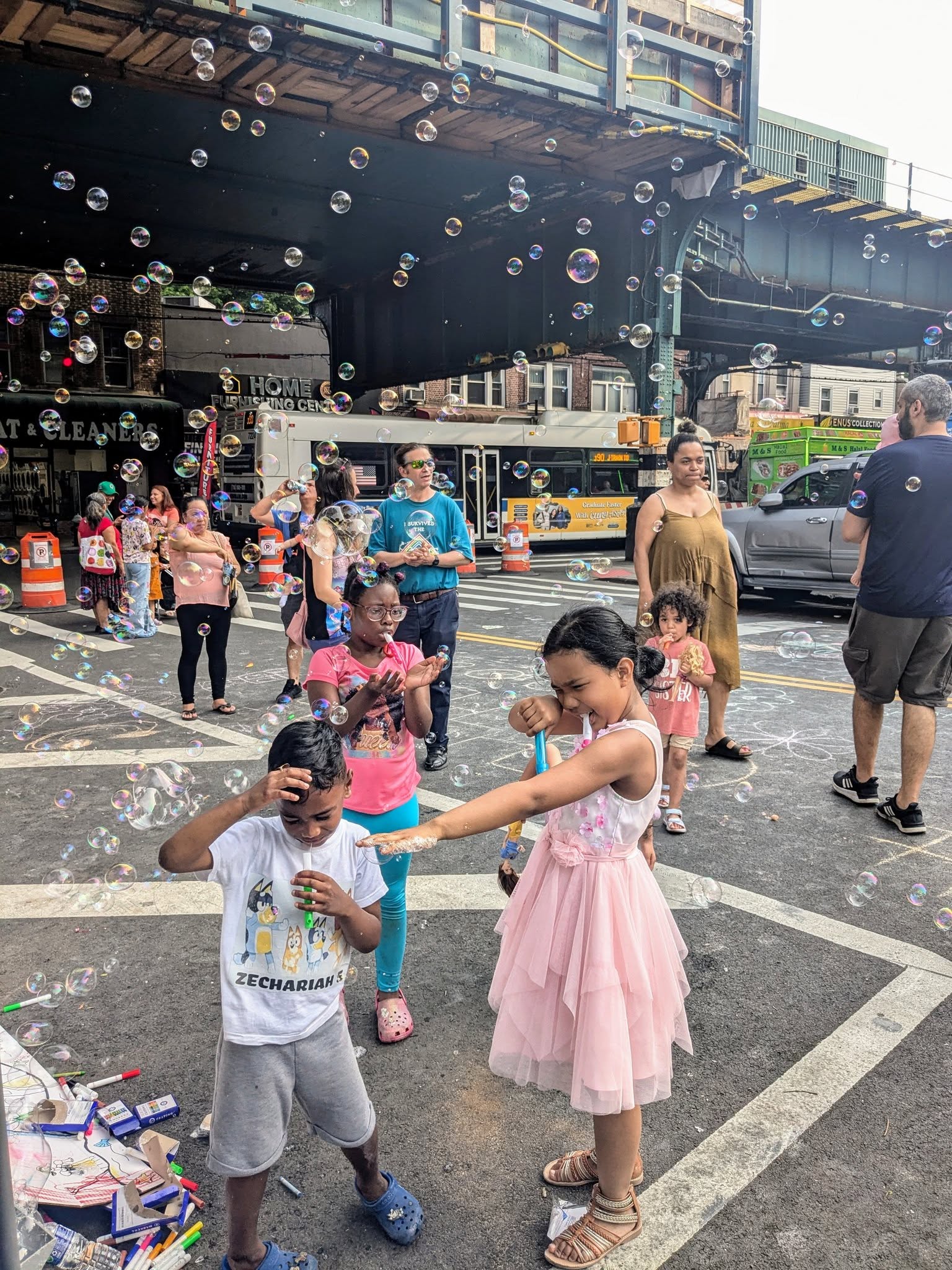 Children and adults enjoy a lively street scene filled with bubbles. A girl in a pink dress playfully points a bubble wand at a smiling boy, while others watch and play under an elevated train track on a sunny day.