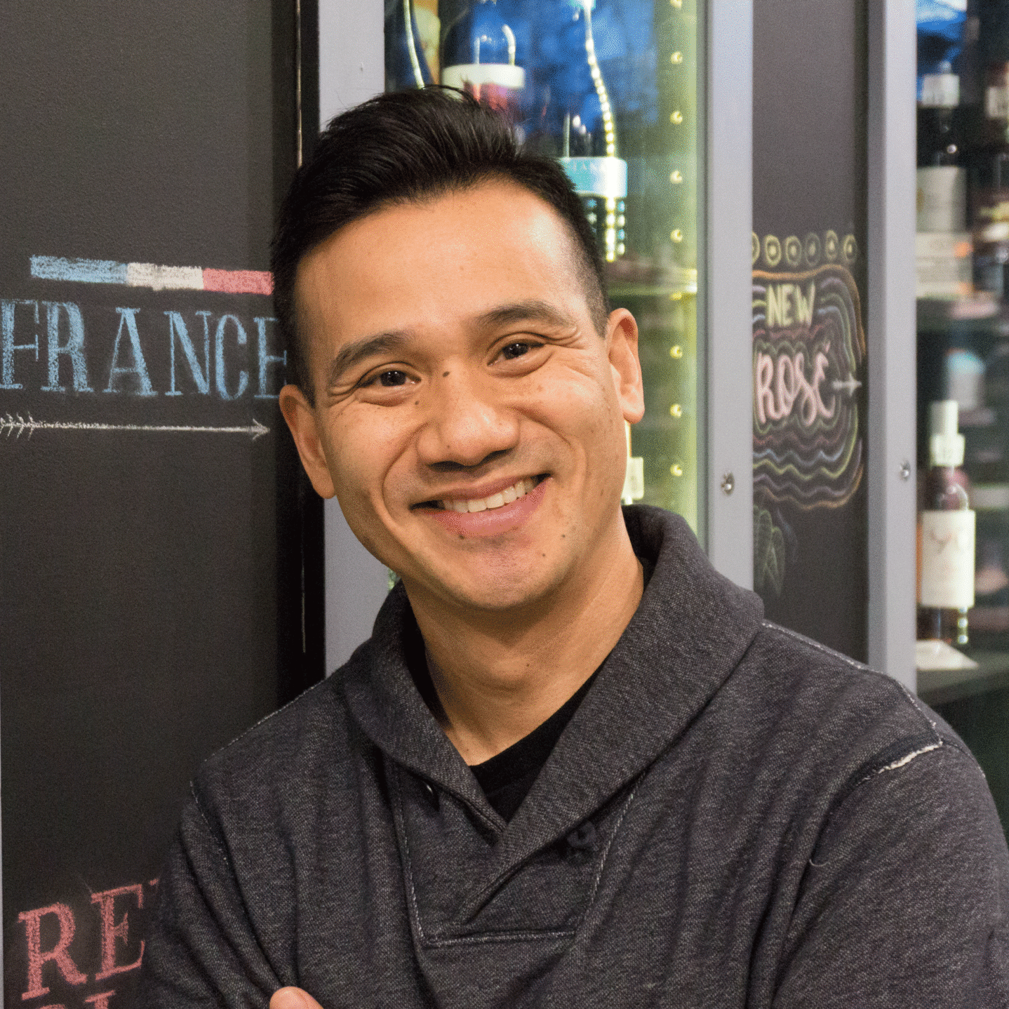A man with short dark hair smiles and stands with arms crossed in front of a display of wine bottles. Shelves and bottles fill the background, and ``France`` is written on a blackboard behind him.