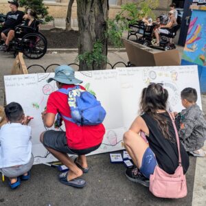 Four children and an adult kneel on a sidewalk, drawing on a large white board with colorful markers. Other people sit nearby under trees and on benches, and a vibrant utility box is visible in the background.