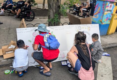 Four children and an adult kneel on a sidewalk, drawing on a large white board with colorful markers. Other people sit nearby under trees and on benches, and a vibrant utility box is visible in the background.