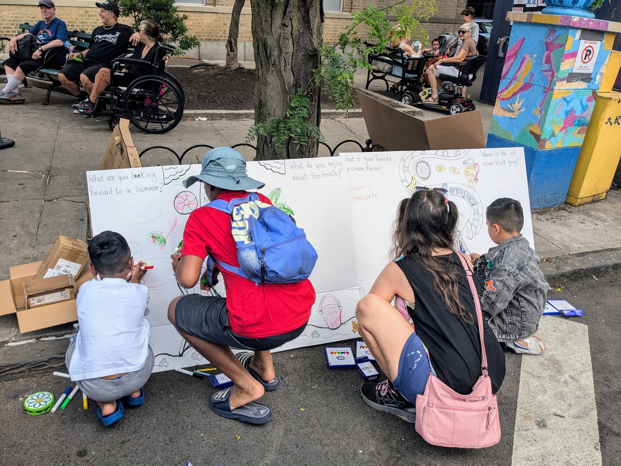 Four children and an adult kneel on a sidewalk, drawing on a large white board with colorful markers. Other people sit nearby under trees and on benches, and a vibrant utility box is visible in the background.
